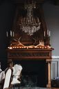 Brown wooden fireplace with antler decor and old fashioned candlesticks on mantelpiece in cozy room with elegant chandelier and fur covered chairs