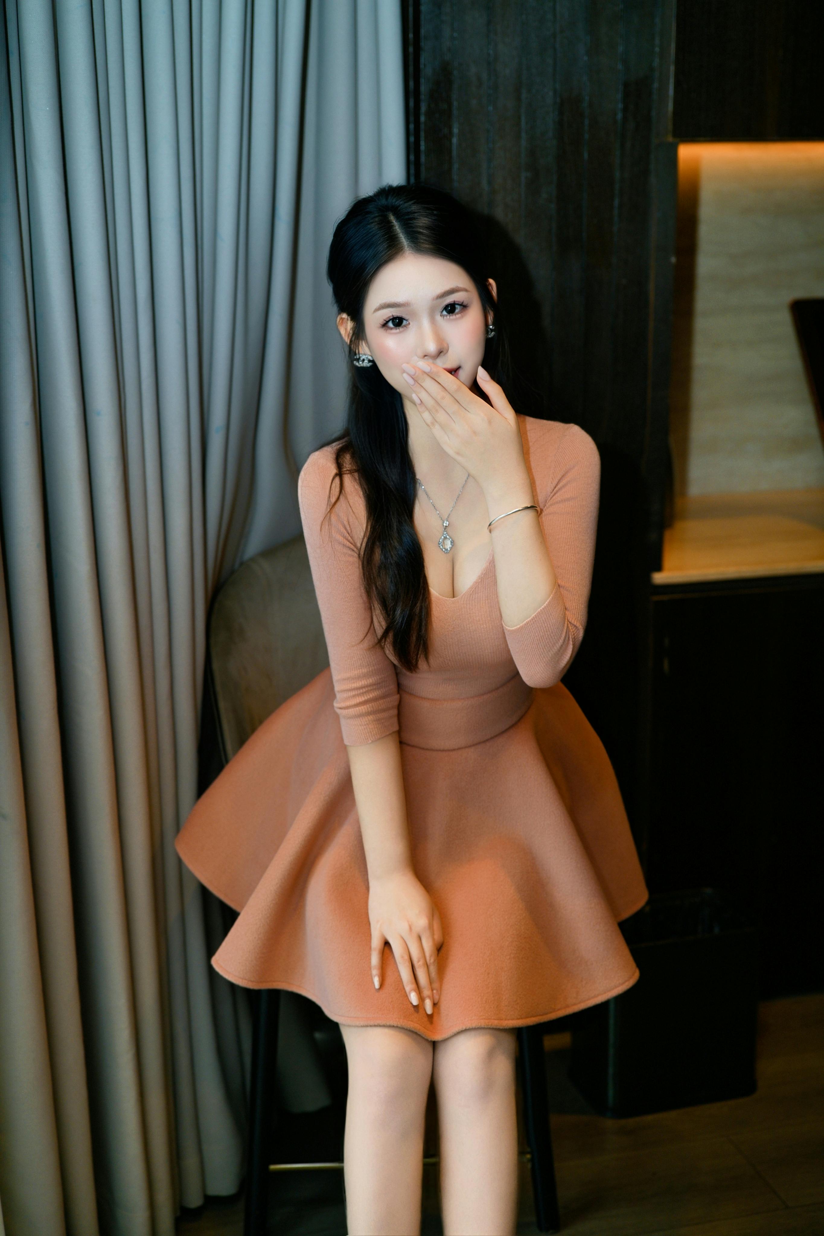 Stylish young woman in a peach dress standing indoors, showcasing elegance and fashion.