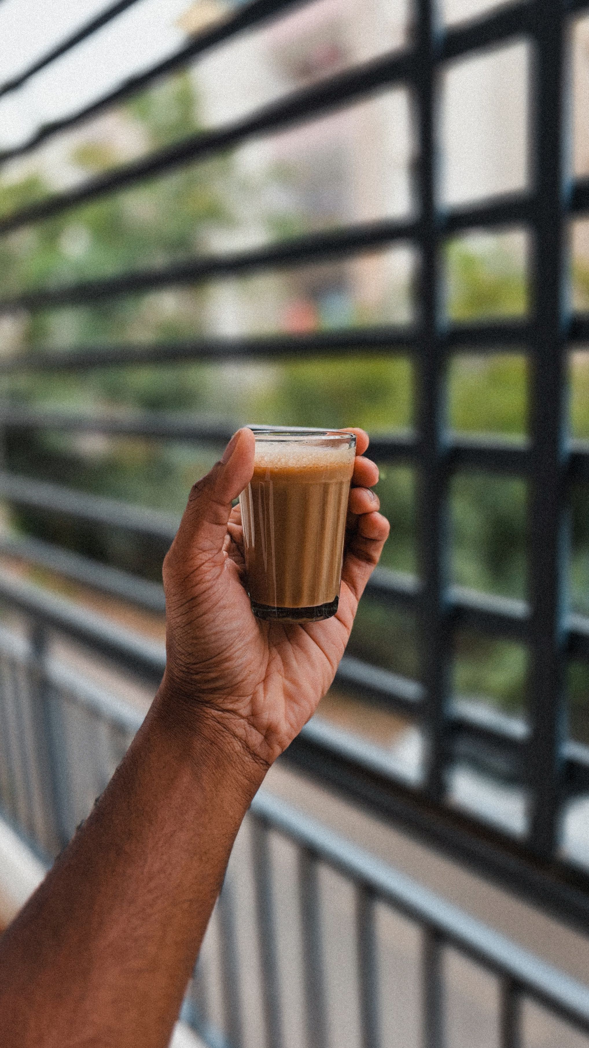 Close-up of a hand holding a glass of morning coffee with a city railing background, evoking a cozy outdoor atmosphere.