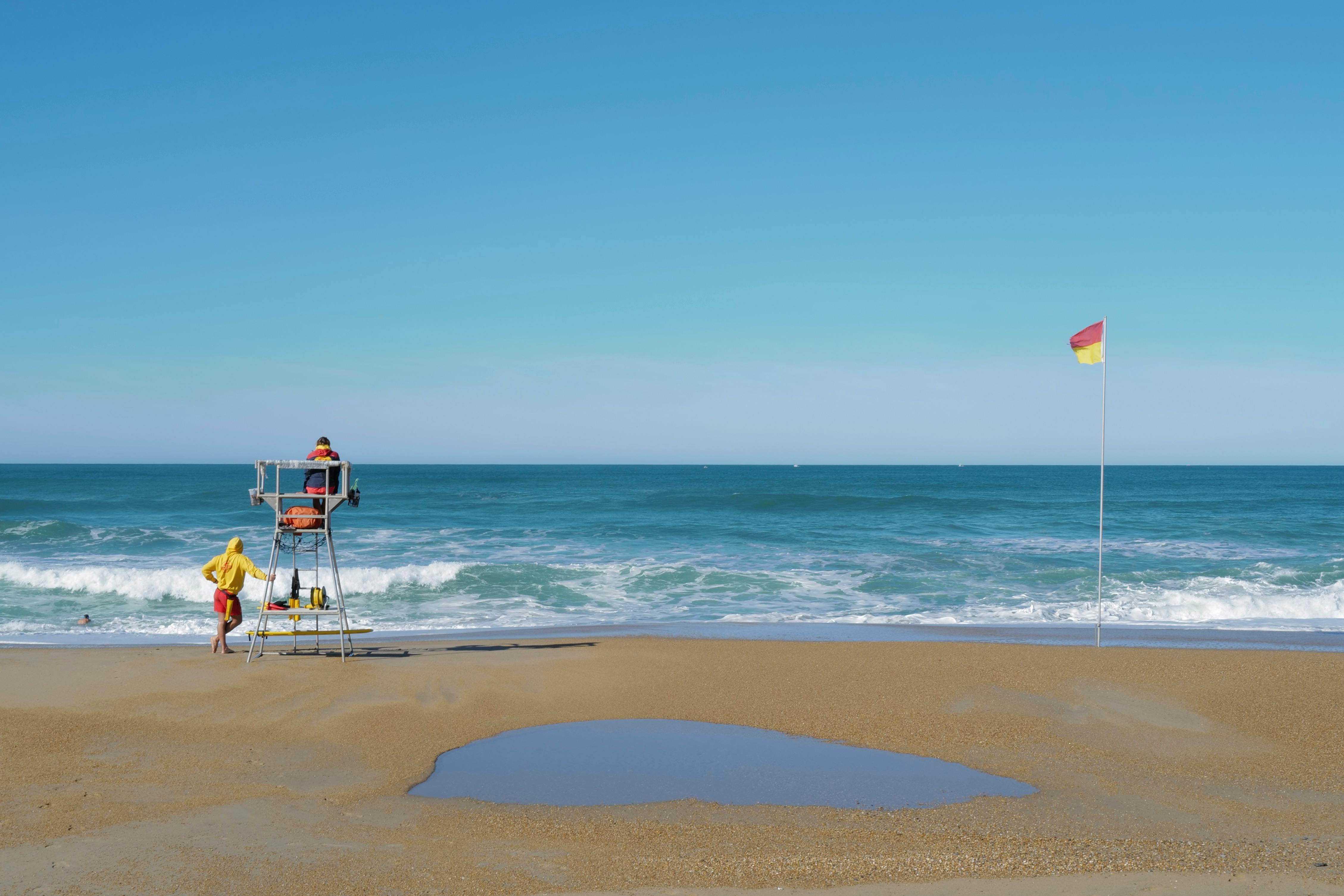 Escena de playa serena con salvavidas, olas y bandera de seguridad bajo un cielo despejado.