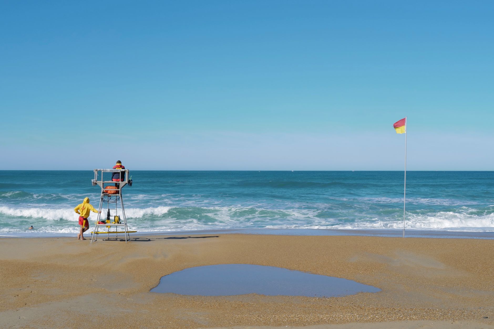Escena de playa serena con salvavidas, olas y bandera de seguridad bajo un cielo despejado.