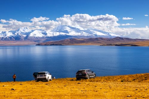 Free A breathtaking landscape showcasing snowy mountains, a serene lake, and parked cars under a clear blue sky. Stock Photo