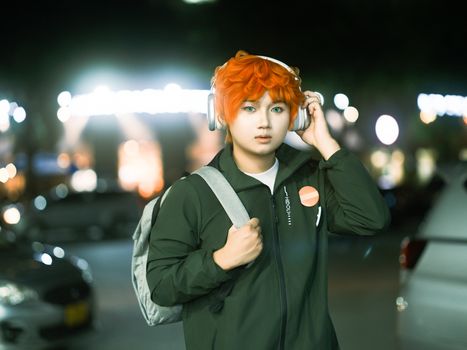 Young adult with striking orange hair and headphones in a nighttime urban setting.