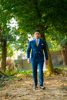 A young man confidently walks in a lush green outdoor setting, wearing a stylish blue suit.