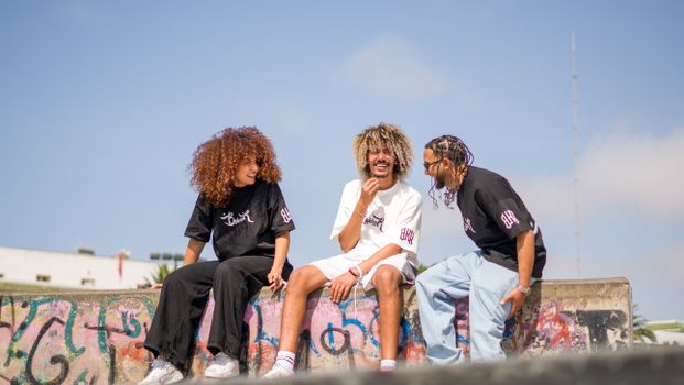 Three young adults sitting on graffiti-covered ramp in skate park, laughing and chatting on a sunny day.