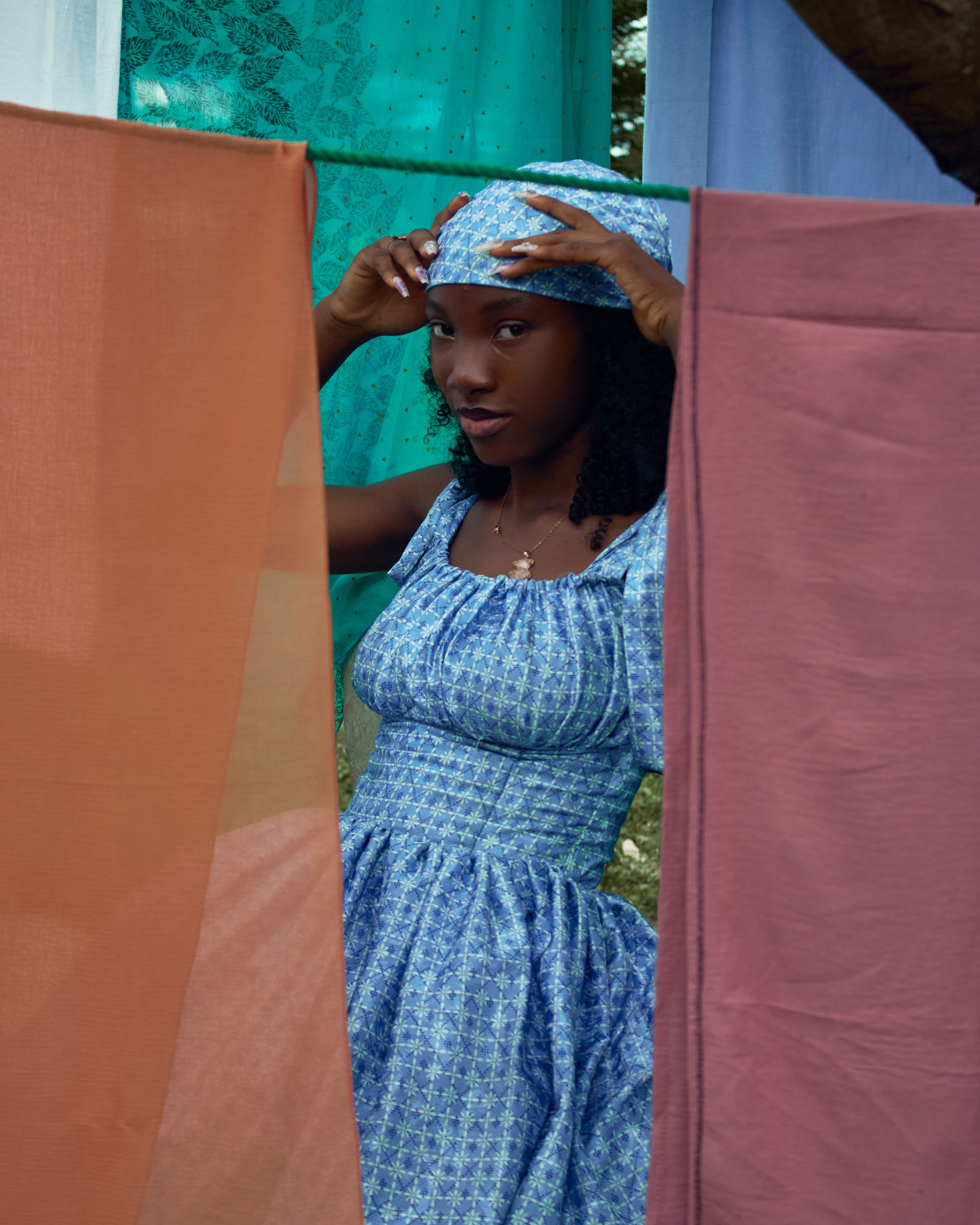 A woman in a blue dress adjusts her headscarf among colorful fabrics outdoors.
