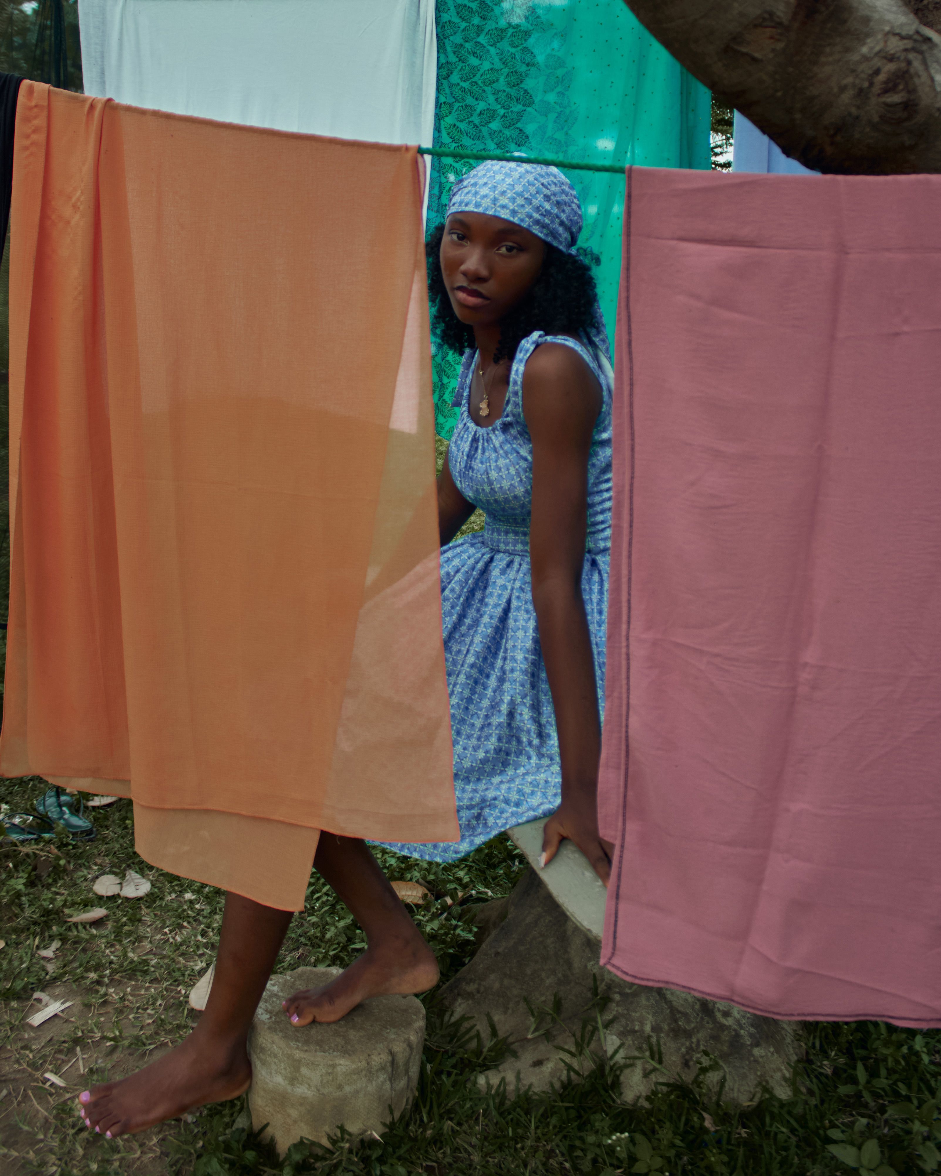 A young woman in a blue dress sits among colorful hanging fabrics outdoors.