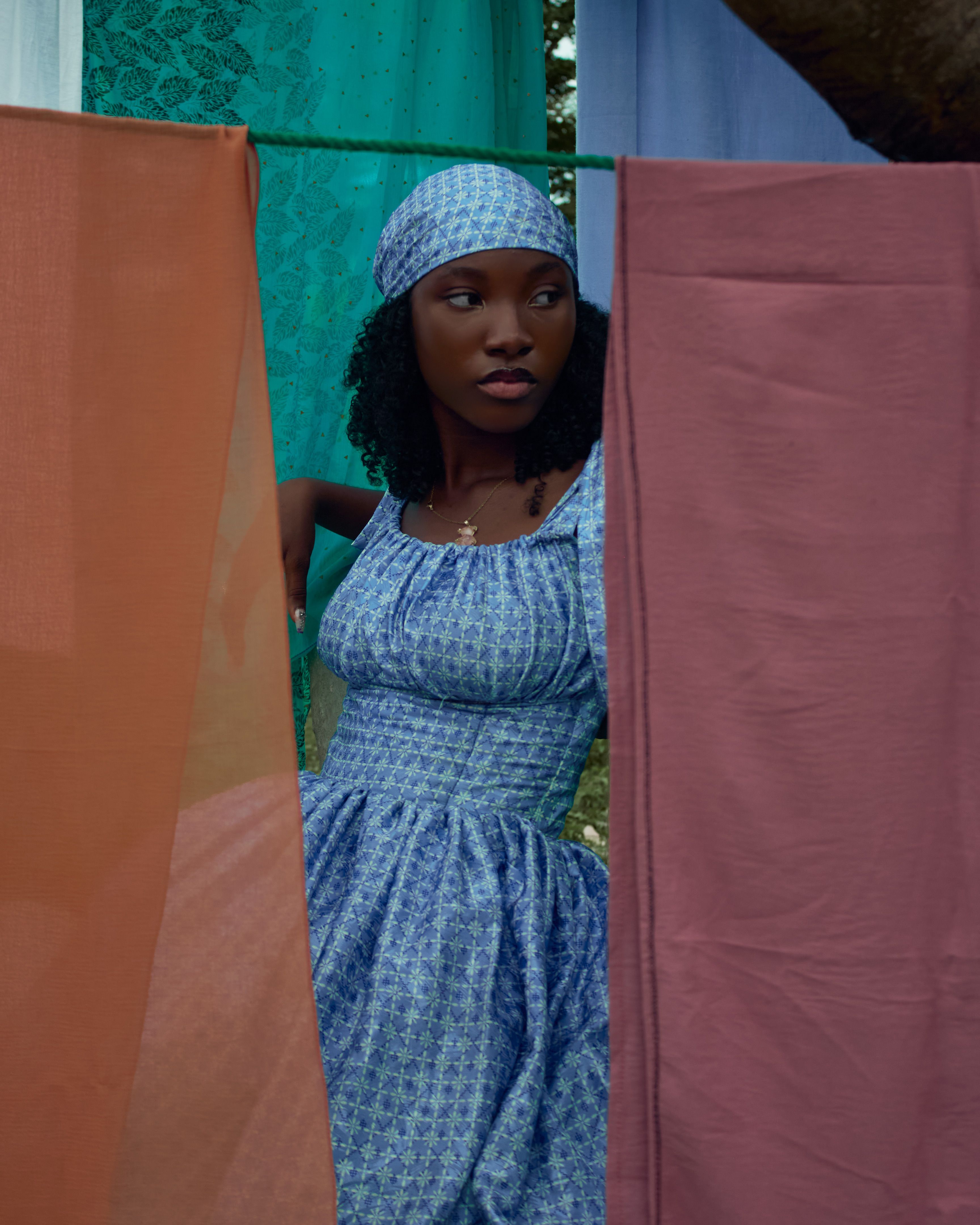 Stylish woman in a blue dress stands between colorful hanging fabrics outdoors.