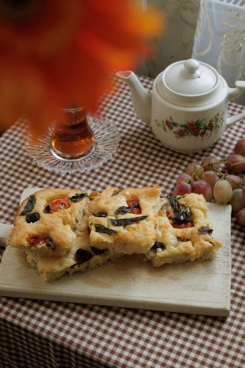 Free Cozy indoor scene featuring homemade focaccia bread with black olives and tomatoes, tea, and grapes on a gingham tablecloth. Stock Photo