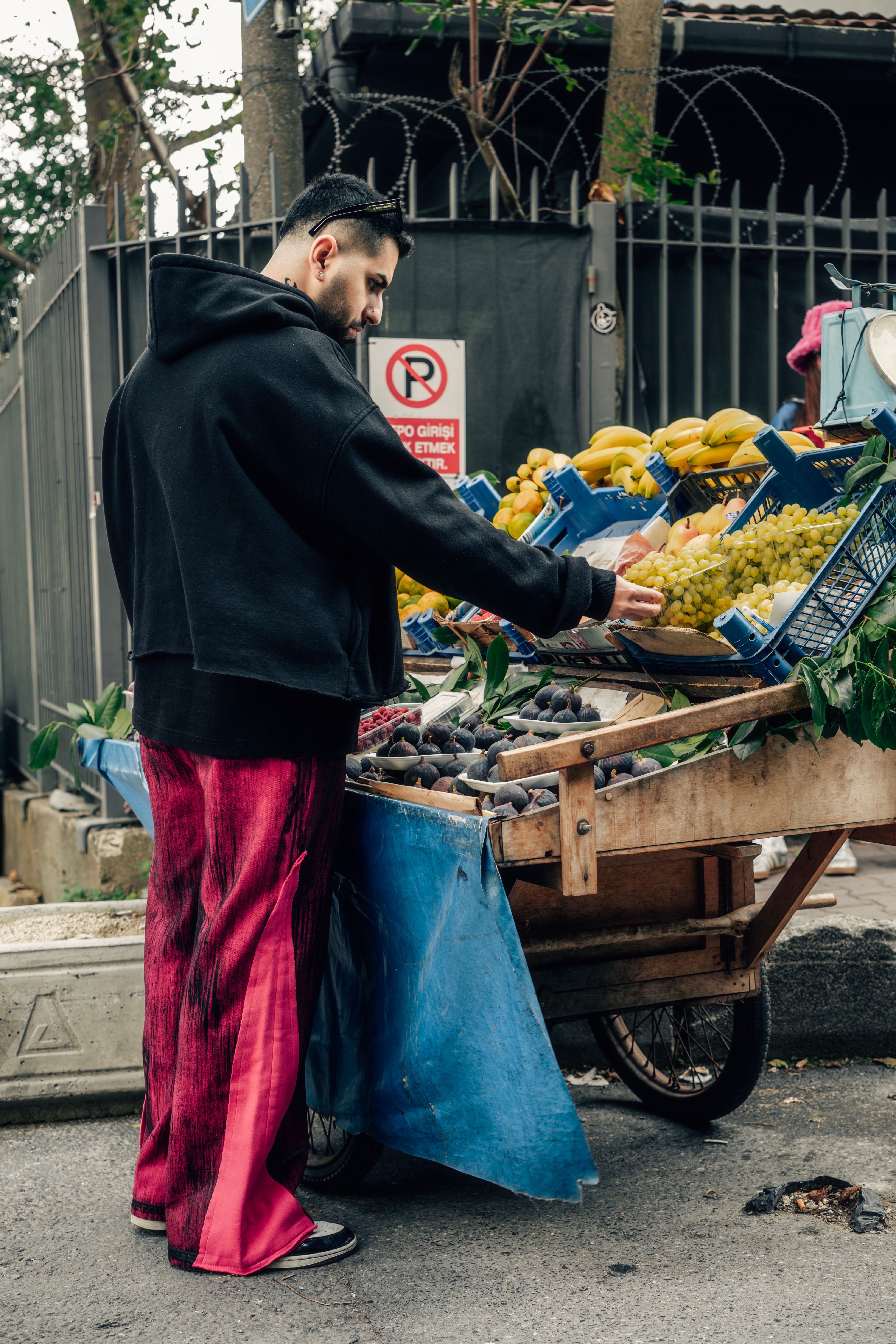 A man in colorful pants selects fruit from a street vendor's cart outdoors.