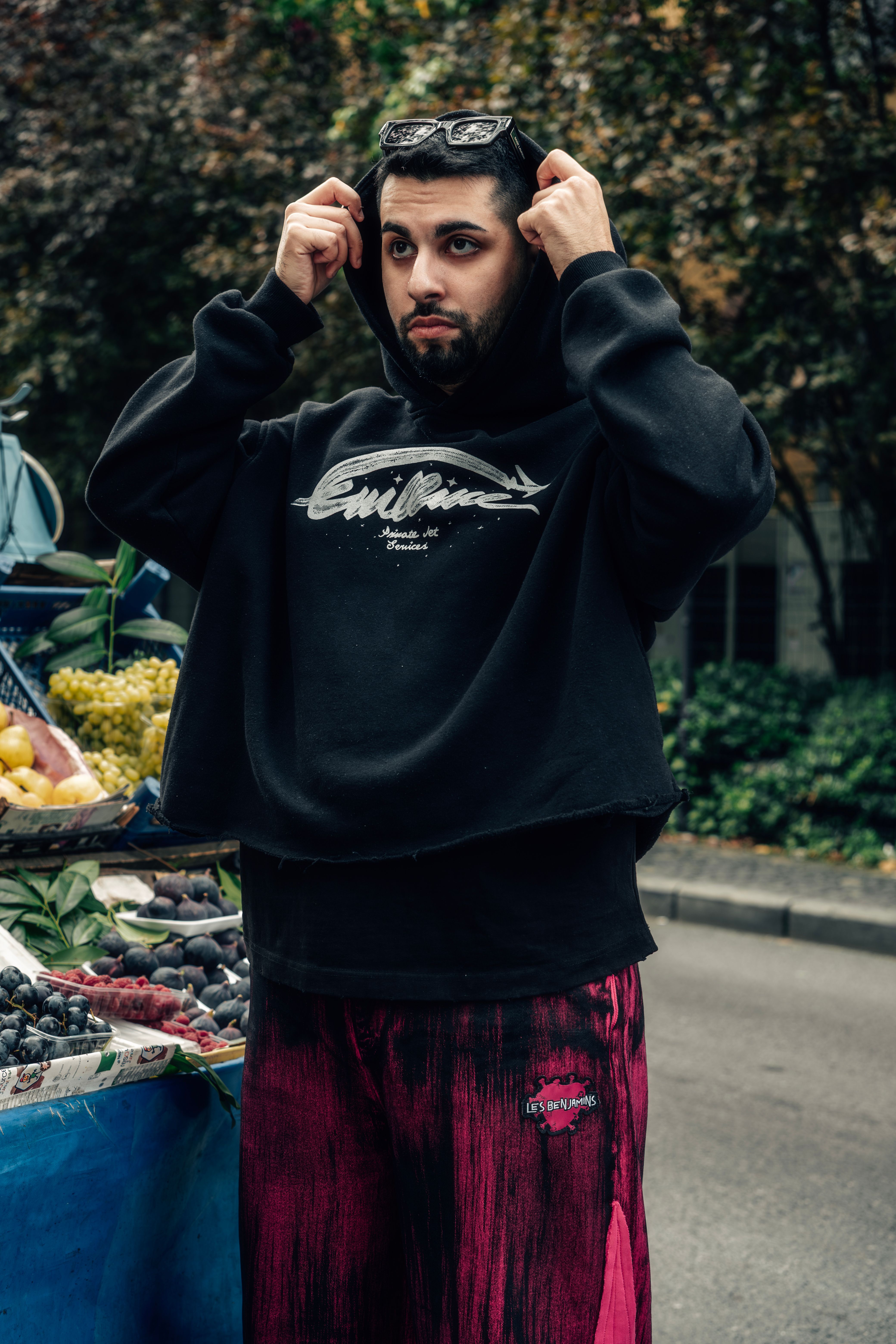 Man wearing hoodie and sunglasses at outdoor fruit market stand.