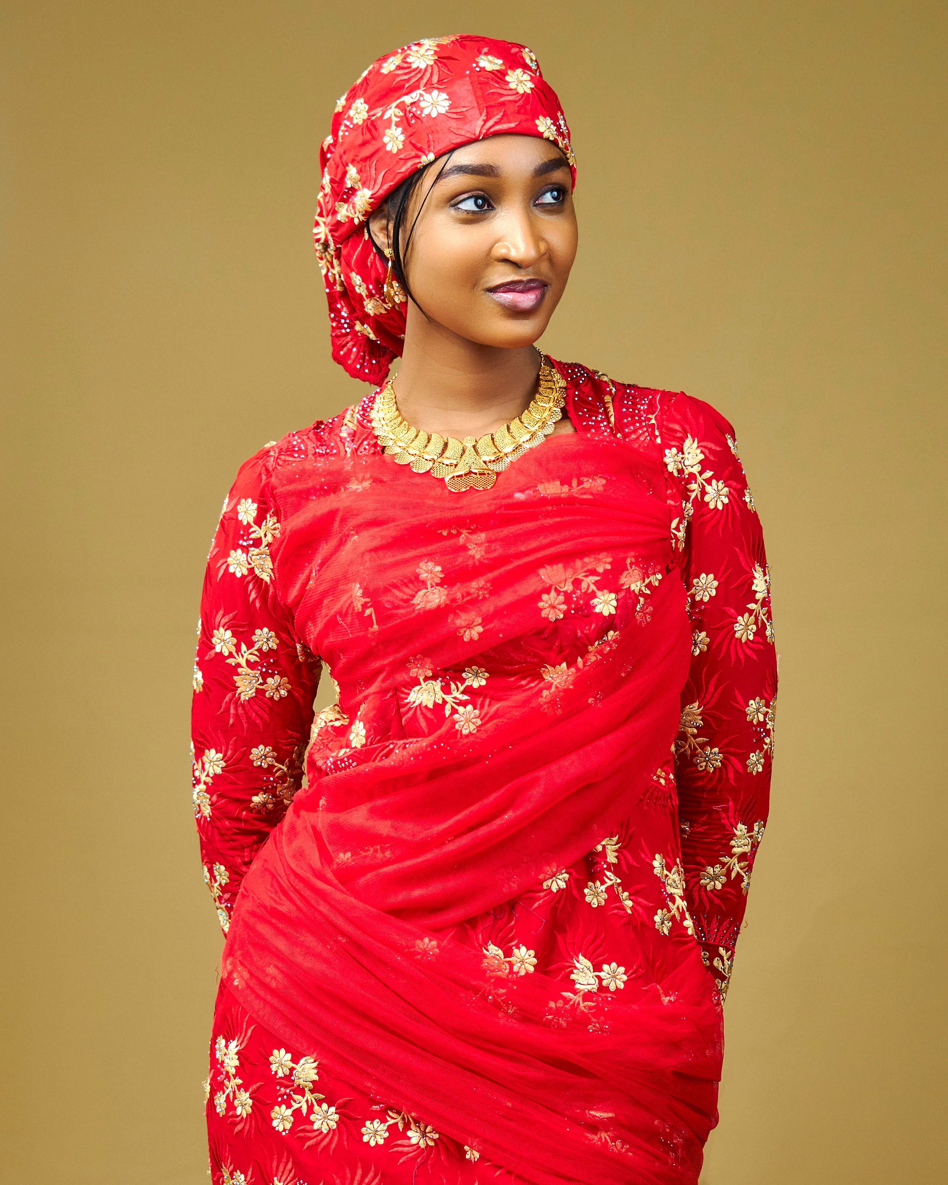 Portrait of a woman in vibrant red traditional dress with gold ornaments against a plain background.