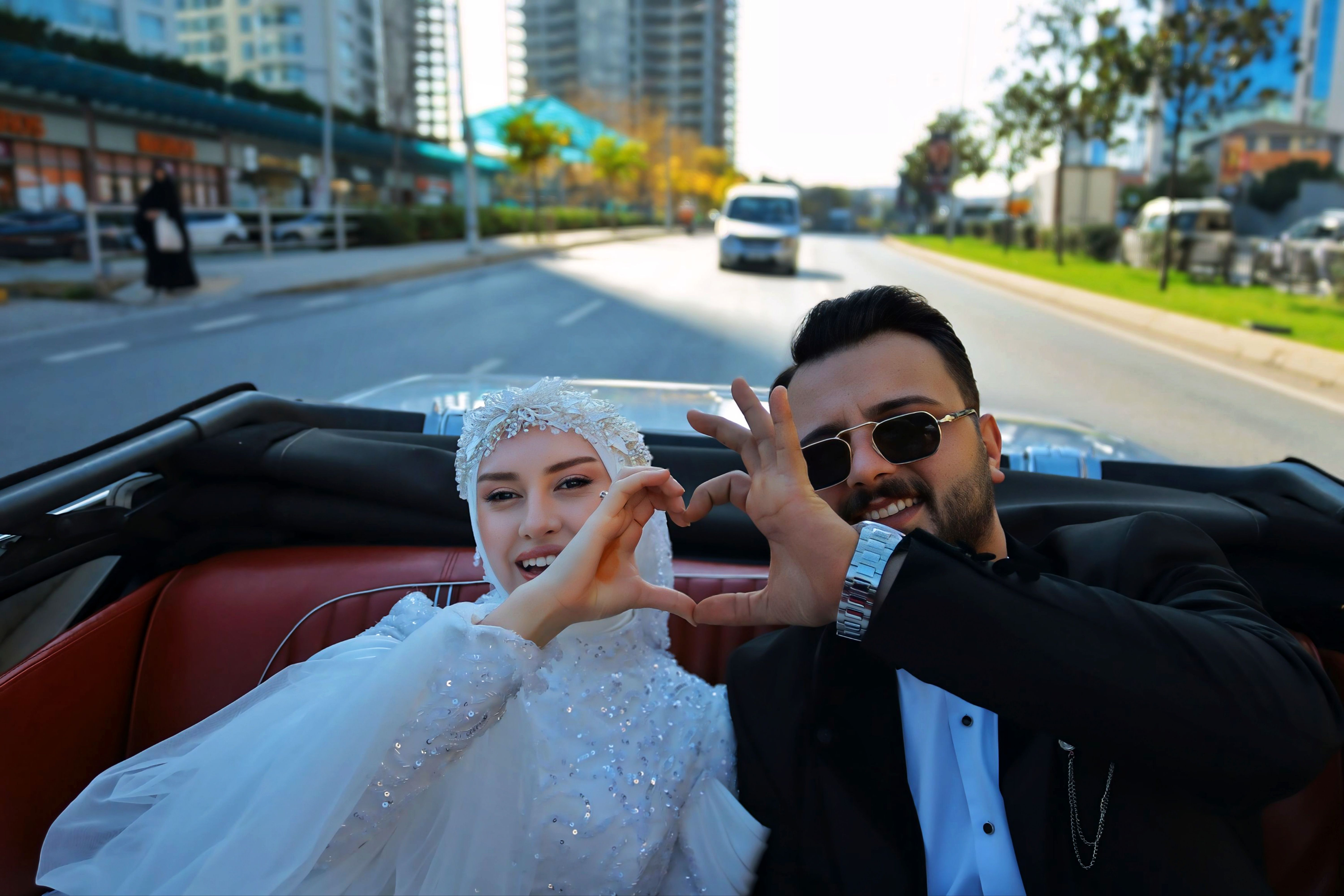 Happy couple in a convertible making a heart shape with hands on a sunny urban street.