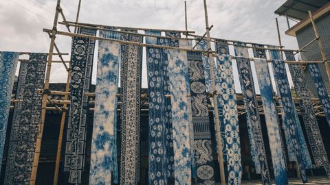 Various indigo dyed fabrics hanging on bamboo racks under the sky, showcasing traditional designs.