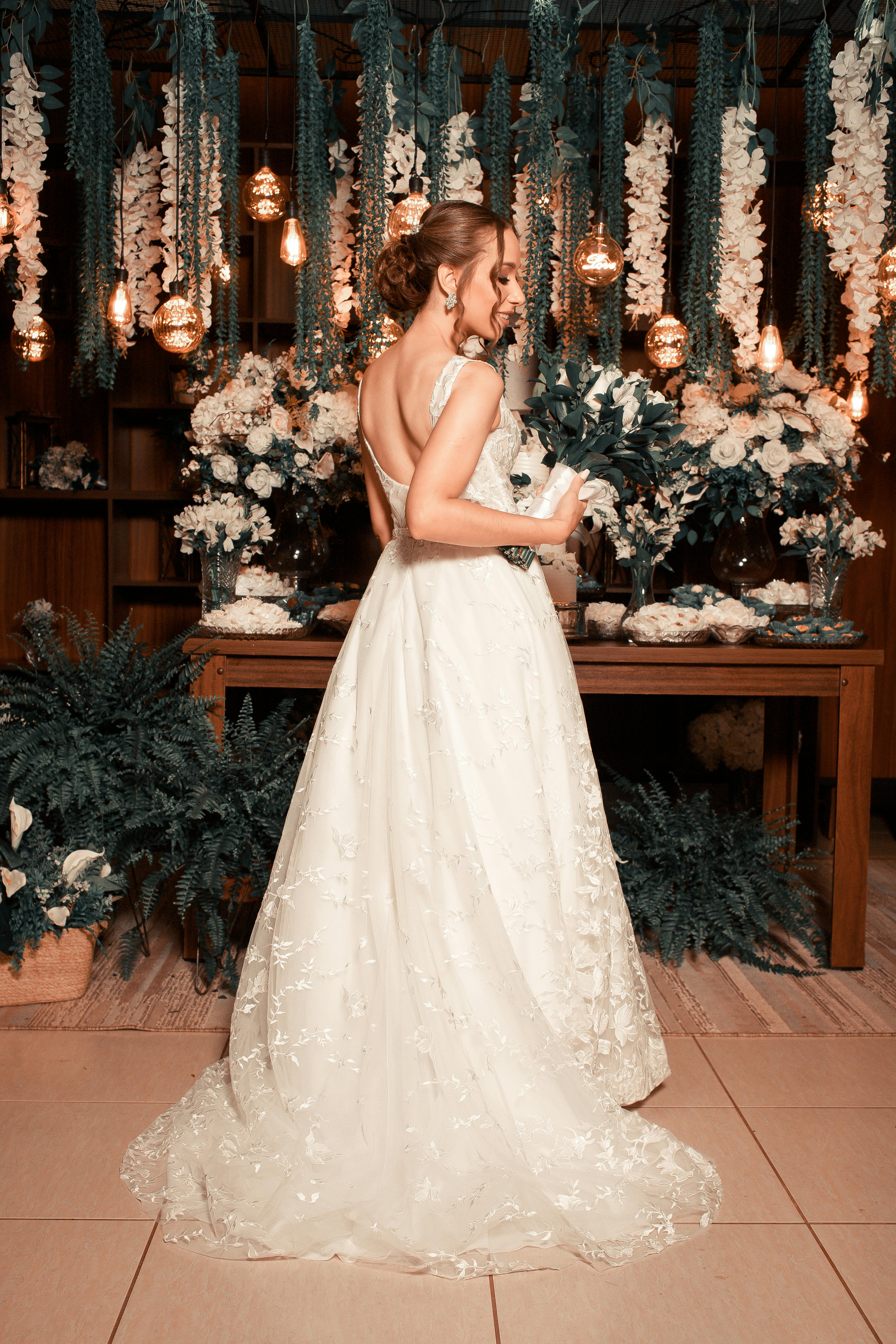 Bride in a lace gown poses with bouquet against floral decor indoors.