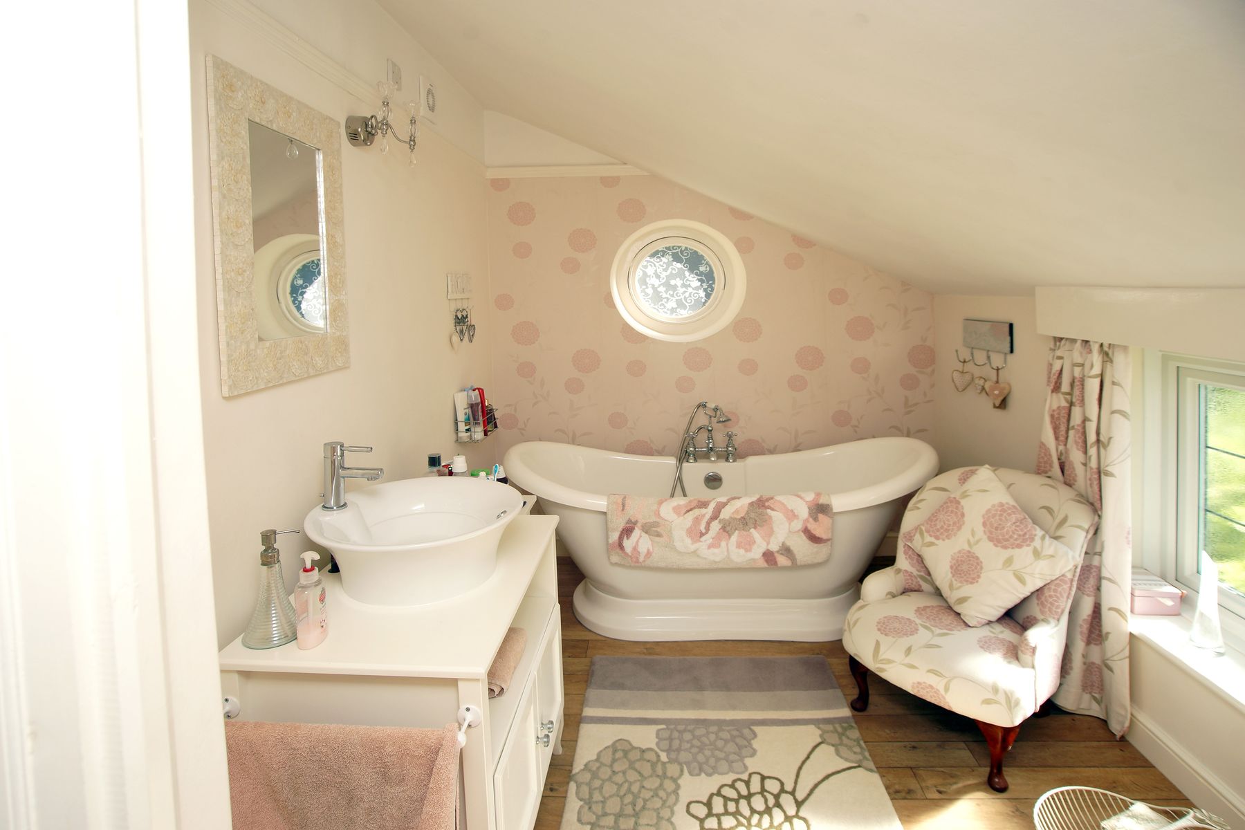 Light-filled attic bathroom with a freestanding white bath, pale wood vanity, circular window and restrained floral details