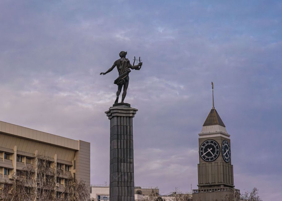 A statue atop a column with a nearby clock tower under a cloudy sky.