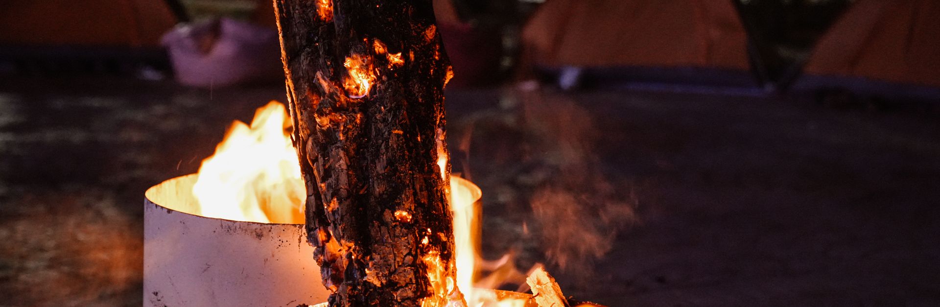Glowing campfire in a barrel among tents, creating a warm nighttime ambiance.