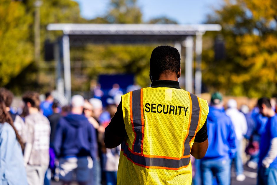 Angry School Security Guard Charged With Making Bomb Threat 2 A security guard monitors a crowd at an outdoor event, ensuring safety and order. | BocaVibe