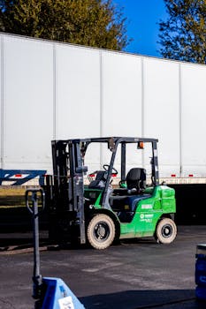 A green forklift parked beside a large white truck outdoors on a clear day.