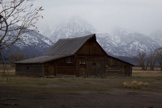 Wooden barn set against mountain range in Wyoming, USA, under overcast sky.