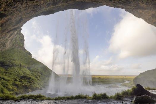 Stunning perspective from behind a waterfall arch capturing lush greenery and open sky.