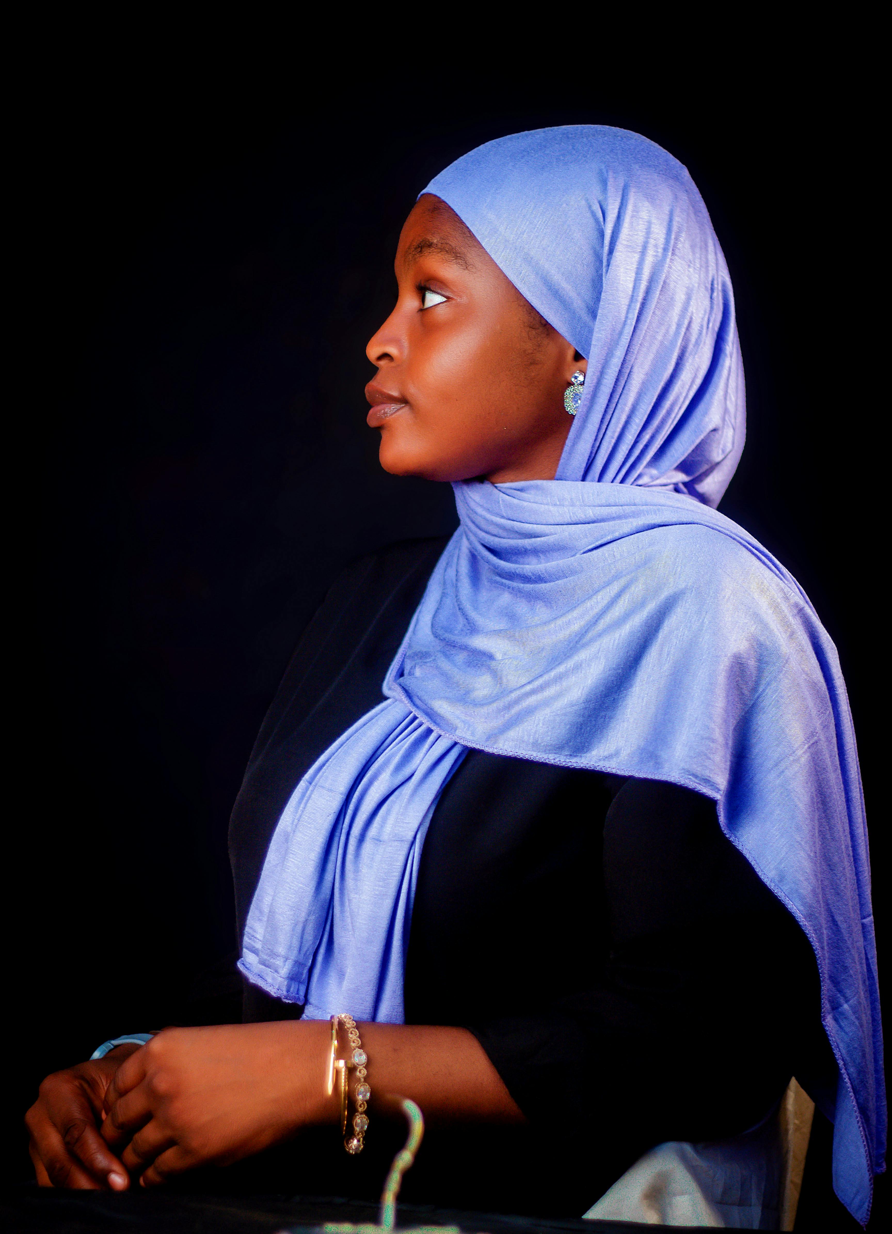 Profile portrait of an elegant woman in a lavender hijab, set against a dark background.