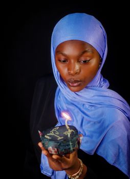 A young woman wearing a blue hijab blows out a candle on a birthday cake.