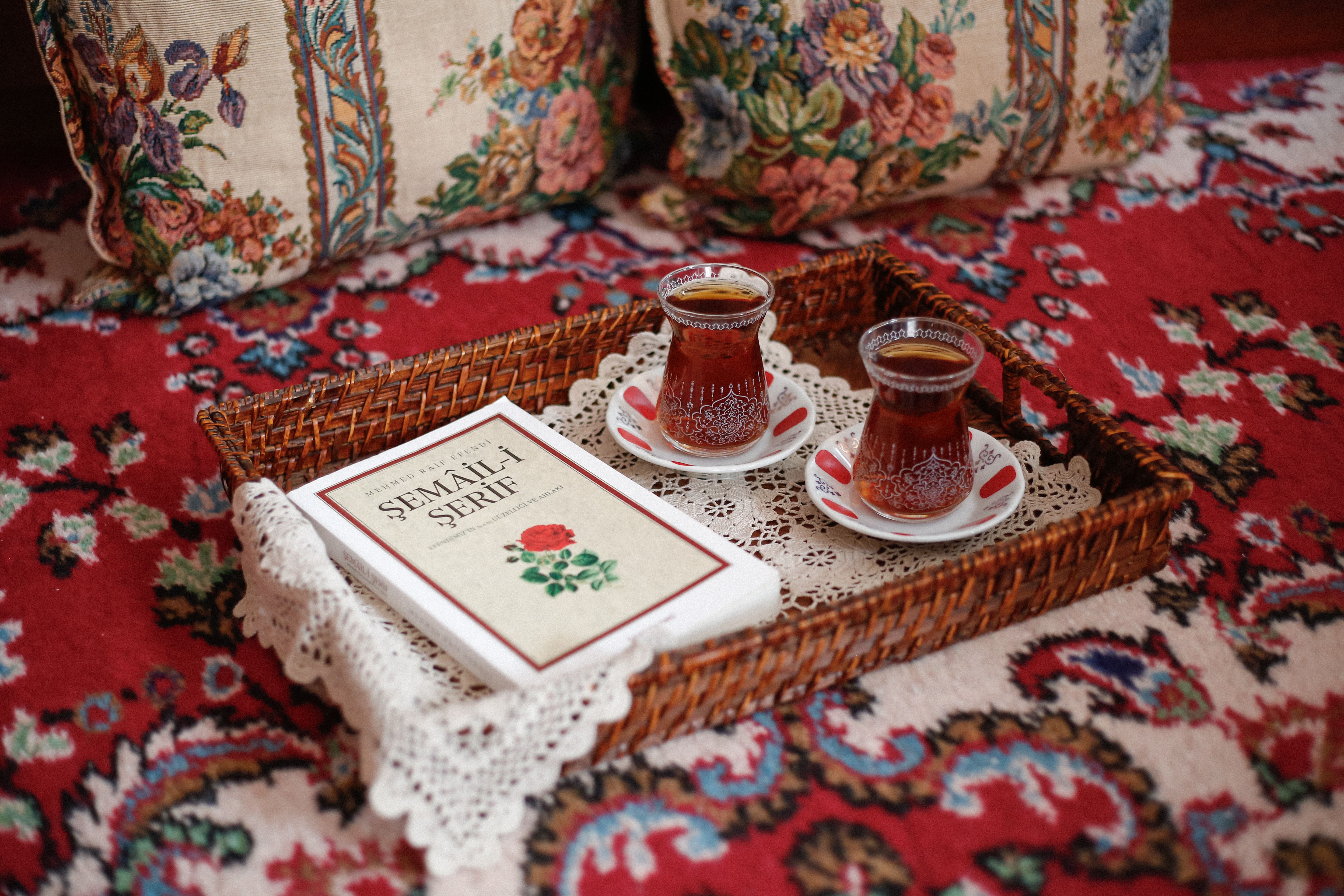 Wicker tray with Turkish tea glasses and book on floral cushions and carpet.