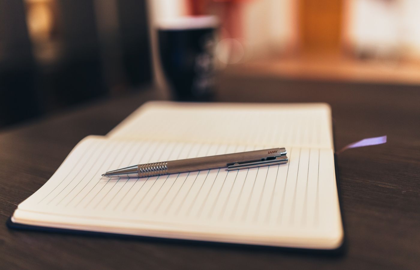 Open notebook with pen on a table, mug in background, cozy setting.
