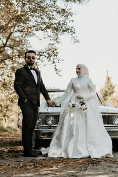 Bride and groom pose elegantly outdoors beside a vintage car on a sunny day.