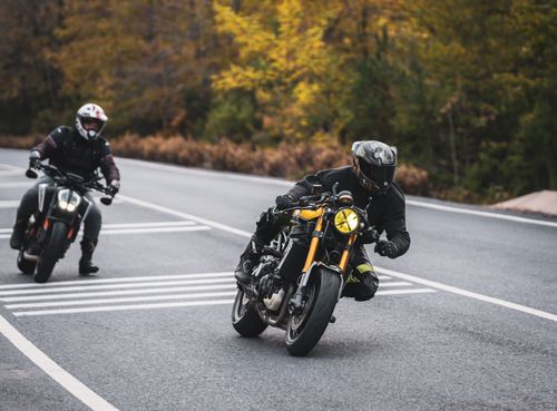 Free Two motorcyclists lean into a curve on a scenic road with autumn foliage in the background. Stock Photo