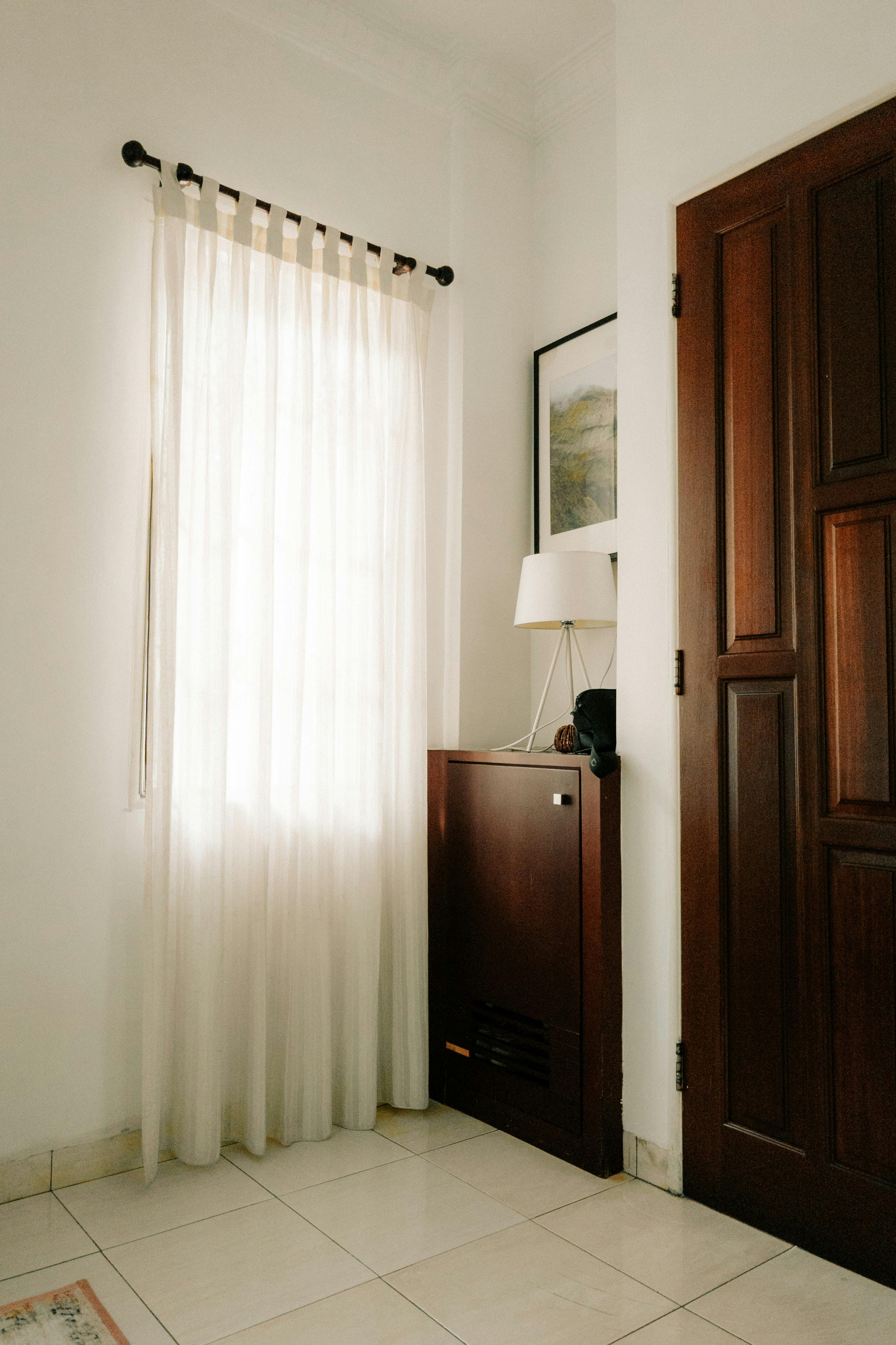 A well-lit corner of a room featuring a wooden door, a lamp, and sheer curtains in a minimalist style.