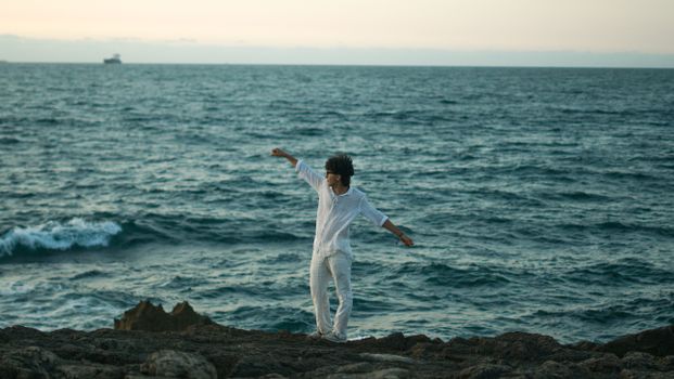 A person in white clothes joyfully dancing on a rocky coast with the ocean in the background during sunset.