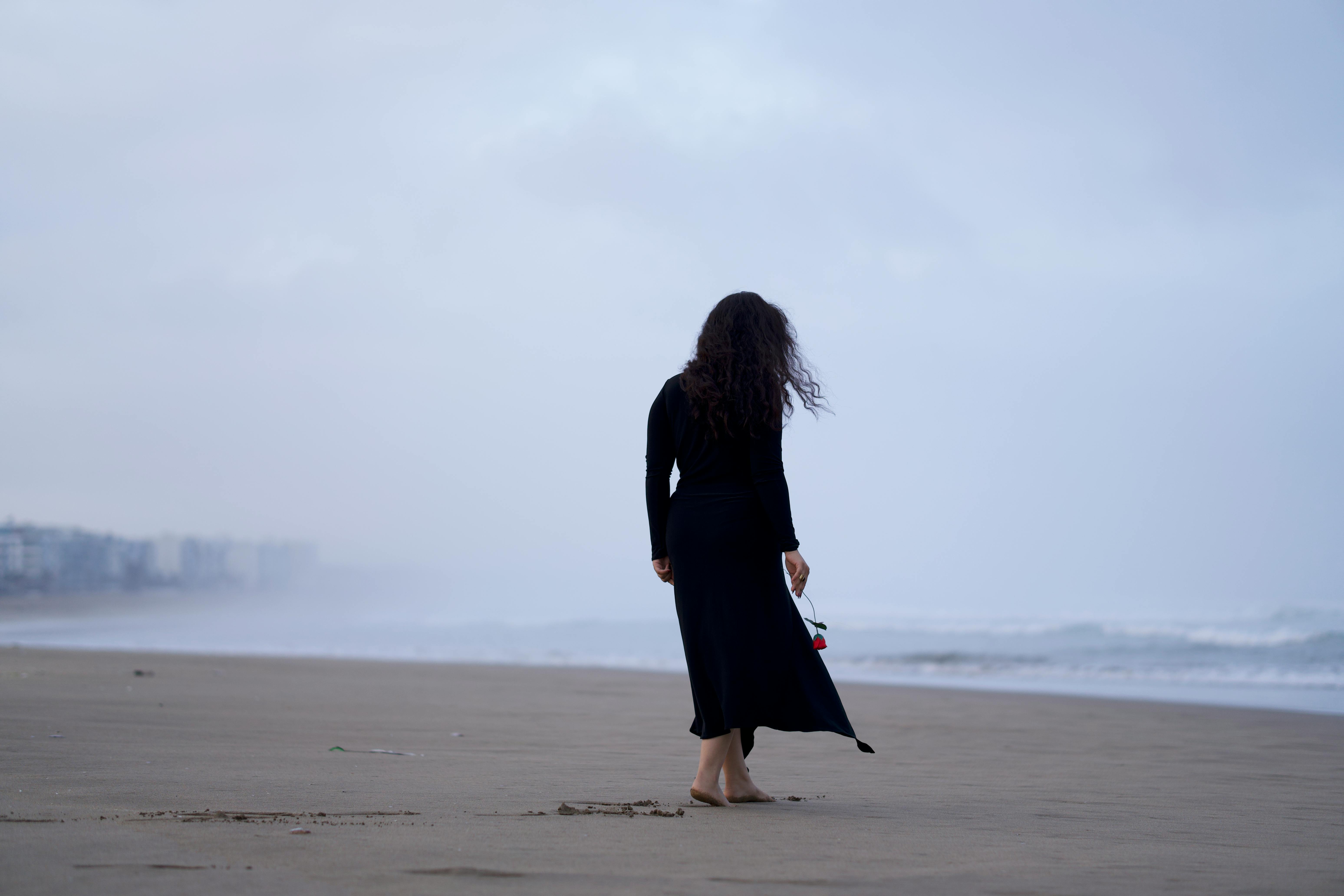 A woman in a black dress walks barefoot on a misty beach holding a flower, conveying solitude.