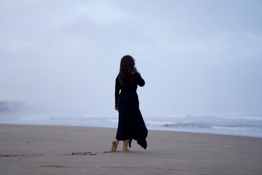 A woman in a black dress with a rose walks alone on a windy beach.