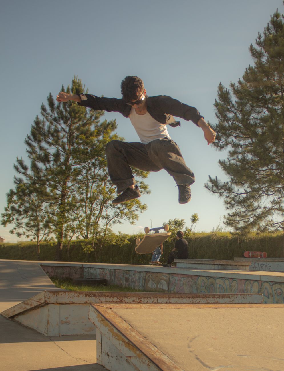 A skateboarder executes a trick mid-air in a sunlit outdoor skate park with trees in the background.. skateboarder, executes,