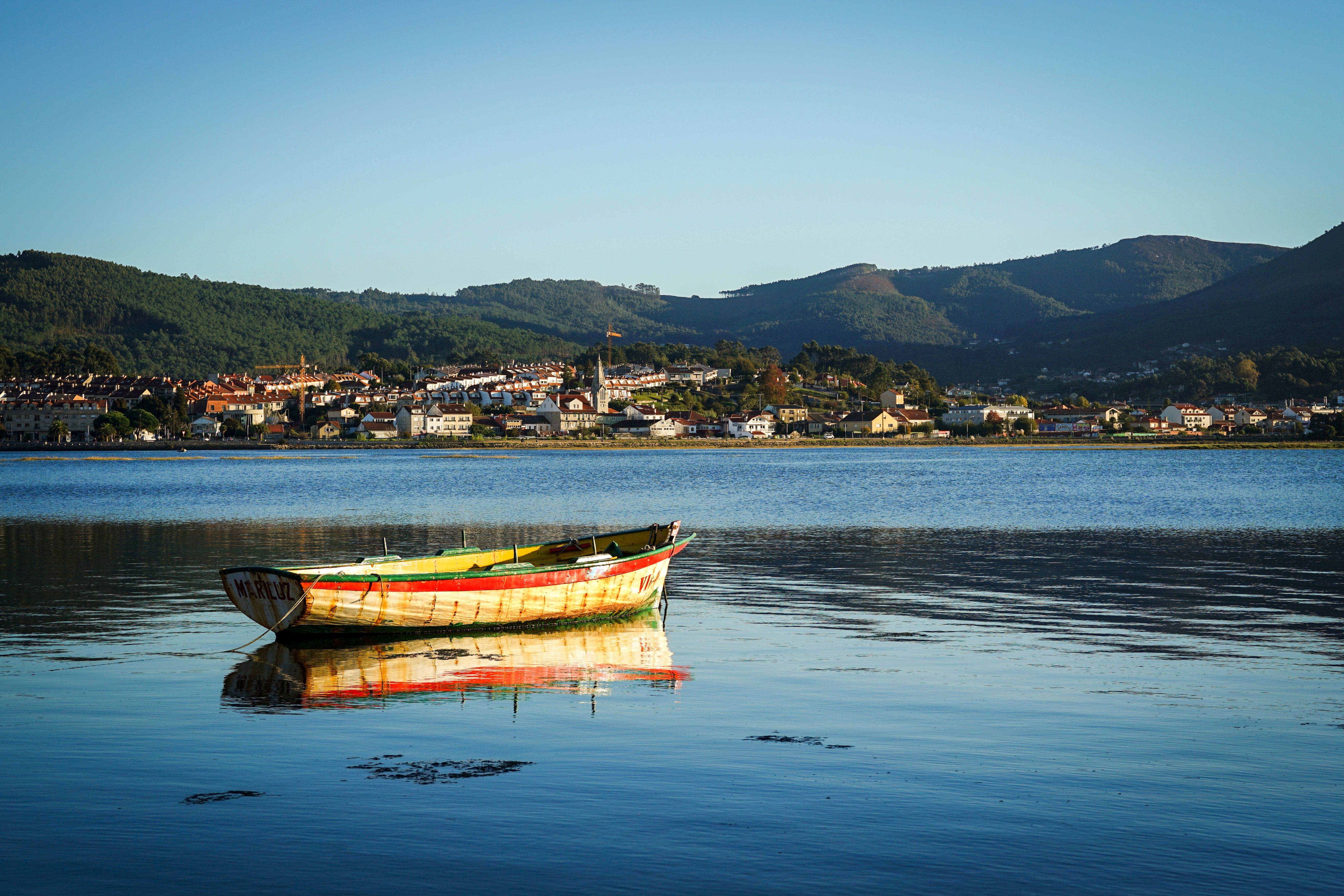 A colorful fishing boat rests on calm waters with a picturesque town and mountains in the background.