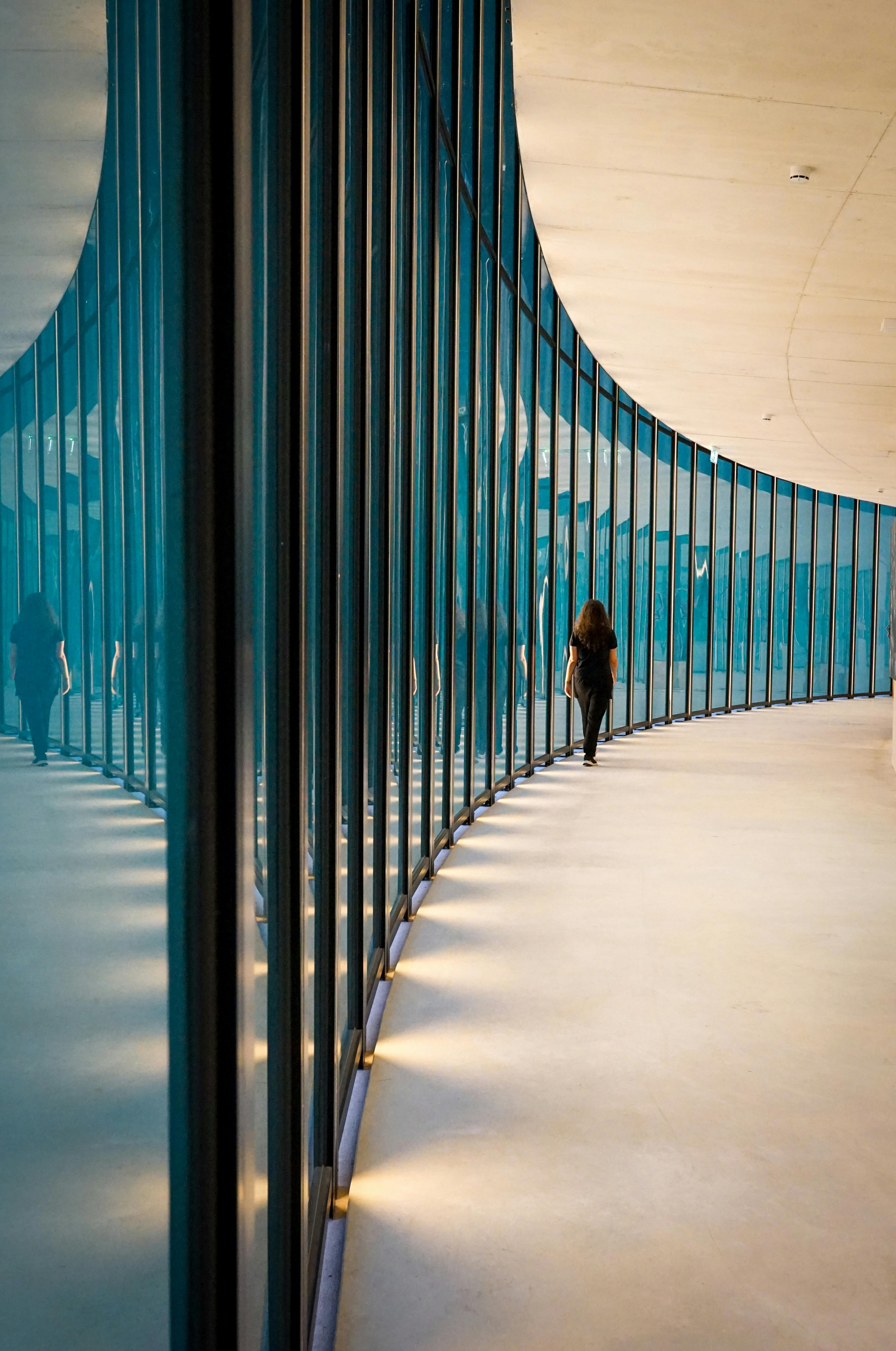 A lone woman walks along a modern curved glass corridor, reflecting an artistic blend of architecture and solitude.