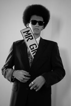 Black and white photo of a man with sunglasses and a sash posing indoors.