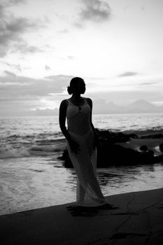 A serene silhouette of a woman standing by the ocean during twilight with gentle waves and distant mountains.