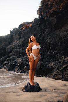 Adult woman in a white bikini posing on a rocky beach during the day. Ideal for travel and lifestyle themes.