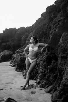 A woman in a bikini poses against rocky cliffs on a beach in this black and white photograph.