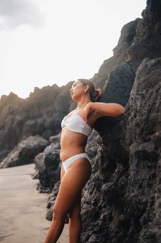 Woman in white bikini leans against rocky cliff at beach during sunset, exuding tranquility and warmth.