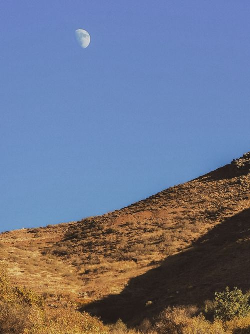 Free A clear blue sky with a visible daytime moon over a rugged mountain landscape. Stock Photo