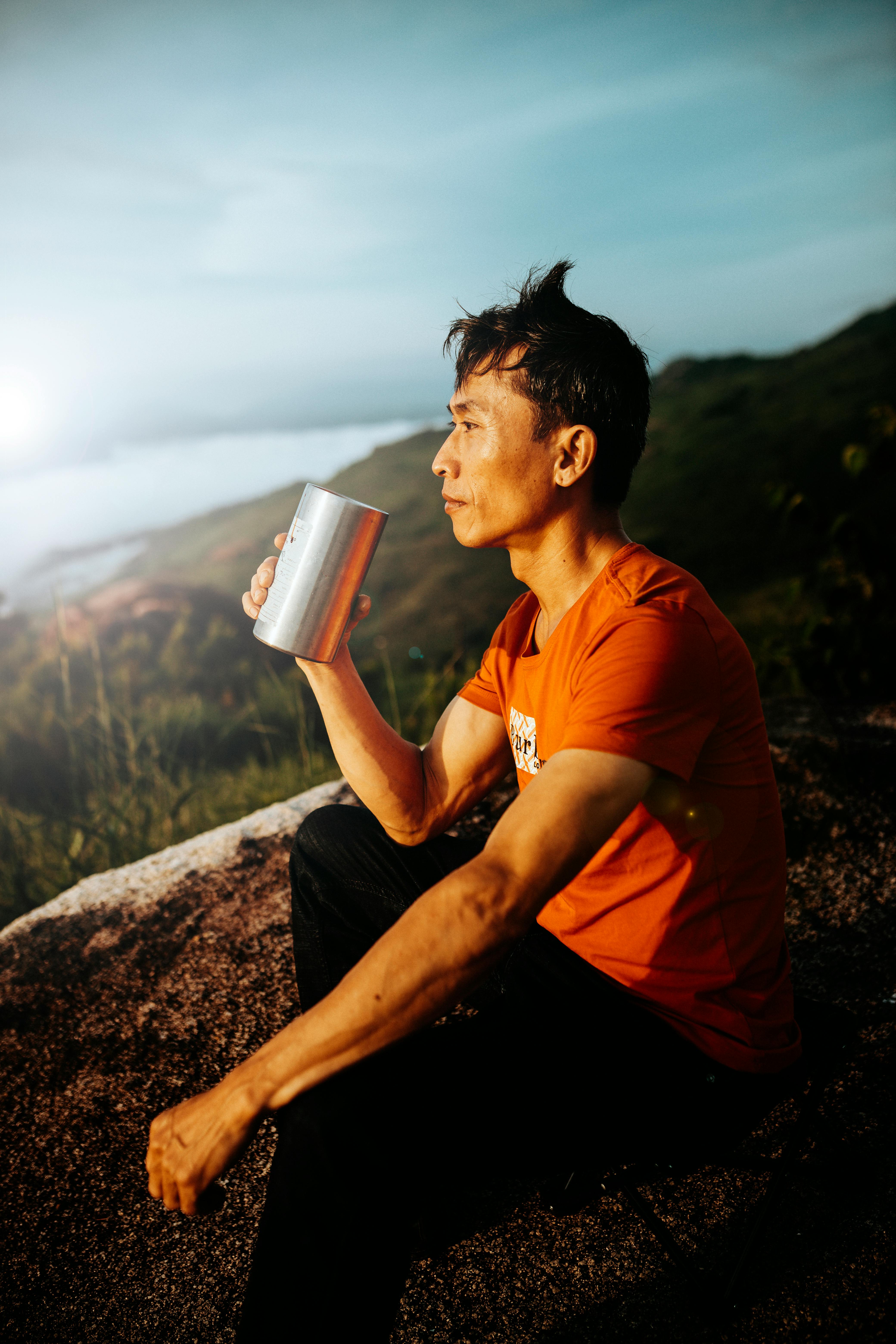 A man in an orange shirt enjoys a cup of coffee on a scenic hill in Kon Tum, Vietnam.