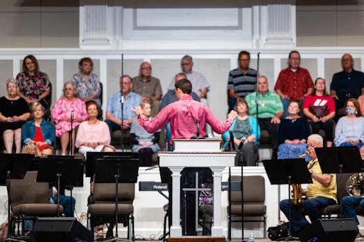 A male conductor leads a choir rehearsal with a group of adults in a church setting.