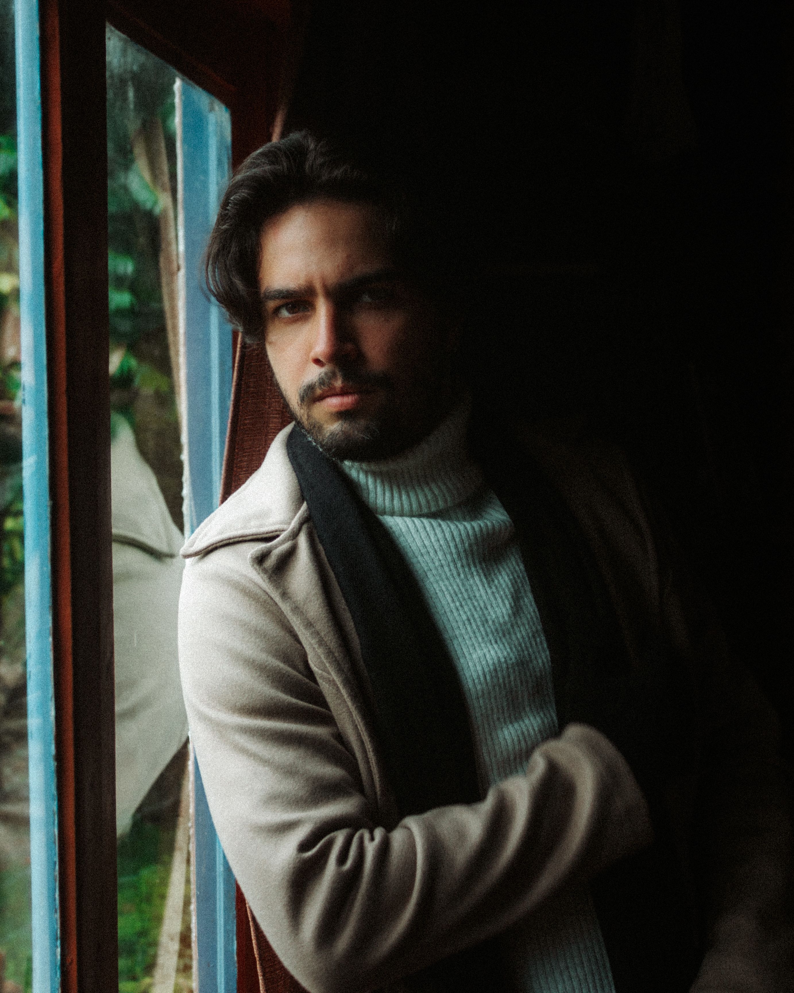 Moody portrait of a man standing by a window dressed warmly, creating a contemplative vibe.