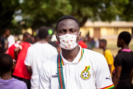 Young man wearing a mask at an outdoor event with a vibrant crowd in the background.