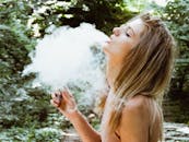 阳光明媚的一天，一位年轻女子在郁郁葱葱的户外环境中吸着电子烟